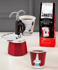Red Moka pot with two cups of coffee and a bag of Bialetti coffee beans on a light surface.