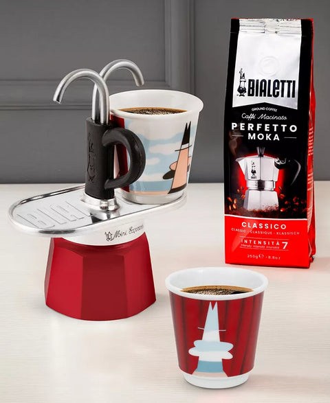 Red Moka pot with two cups of coffee and a bag of Bialetti coffee beans on a light surface.