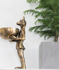 Bronze statue of a figure holding a bowl next to a potted plant on a white background