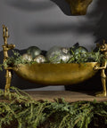Decorative setup with a gold bowl, silver balls, and greenery on a dark background