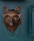 Bronze fox head with glasses on a chain against a teal wall with decorative frames.
