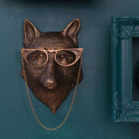 Bronze fox head with glasses on a chain against a teal wall with decorative frames.