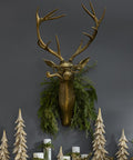 Decorative mantel with gold deer head, greenery, and wooden trees against a dark wall.