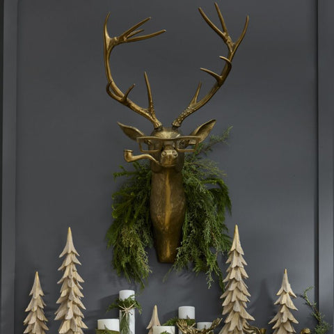 Decorative mantel with gold deer head, greenery, and wooden trees against a dark wall.