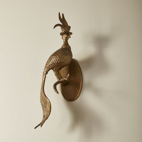 Decorative wall sconce shaped like a peacock on a plain wall with greenery at the bottom.