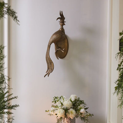 Decorative peacock wall sculpture on a plain wall with a small table and flowers.