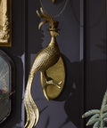 Decorative gold peacock wall sculpture on a dark wall with framed pictures and bottles.