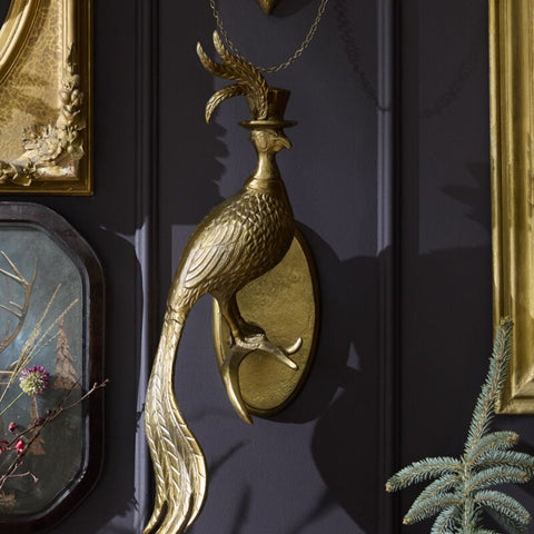 Decorative gold peacock wall sculpture on a dark wall with framed pictures and bottles.