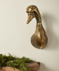 Bronze duck head sculpture on a wooden surface with greenery against a plain wall.