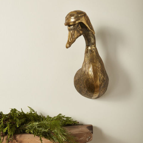 Bronze duck head sculpture on a wooden surface with greenery against a plain wall.