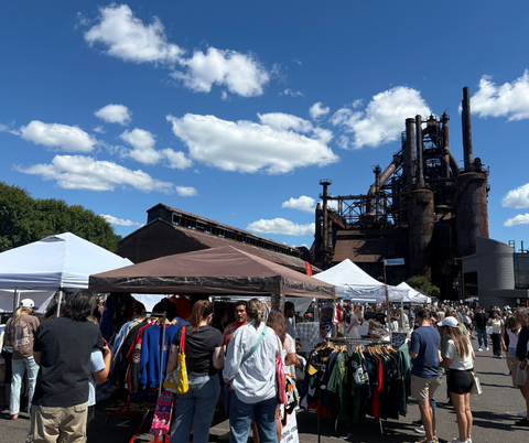 Outdoor market with people and tents against a backdrop of an industrial structure under a blue sky.