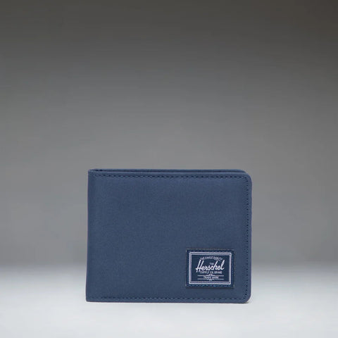 A navy blue bifold wallet with a white Herschel logo patch on the front, set against a dark gray gradient background.