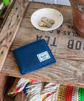 A navy blue leather bifold wallet with a white Herschel logo patch, resting on a rustic wooden crate next to a small bowl of coins and a terracotta pot.