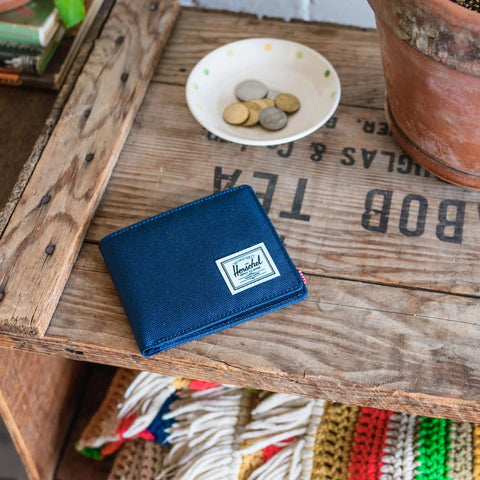 A navy blue leather bifold wallet with a white Herschel logo patch, resting on a rustic wooden crate next to a small bowl of coins and a terracotta pot.