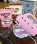 Pink hand mixer with 'SMEG' branding, Old School Candy cookie mix, and oven mitt on a counter.