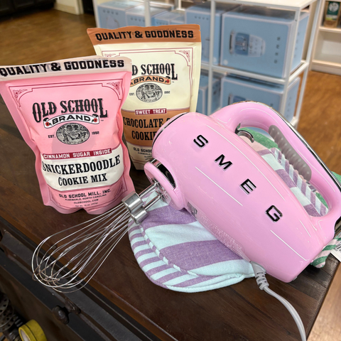 Pink hand mixer with 'SMEG' branding, Old School Candy cookie mix, and oven mitt on a counter.