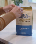 Person holding a coffee bag labeled 'Narn & Hardart' on a kitchen counter with a coffee maker in the background.