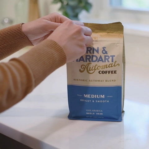 Person holding a coffee bag labeled 'Narn & Hardart' on a kitchen counter with a coffee maker in the background.