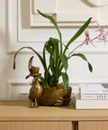 Decorative plant in a gold pot on a wooden surface with books and a framed picture in the background.