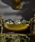 Decorative setup with gold bowl, silver ornaments, and greenery on a dark background